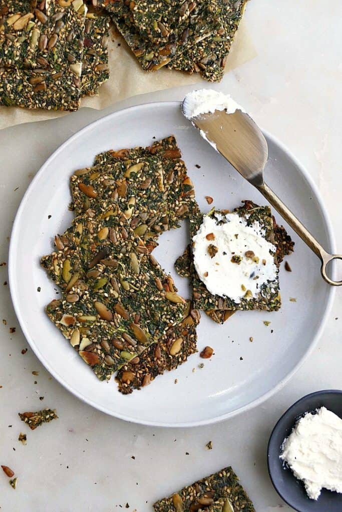 5 seed and kale crackers on a white plate with cream cheese spread on a cracker