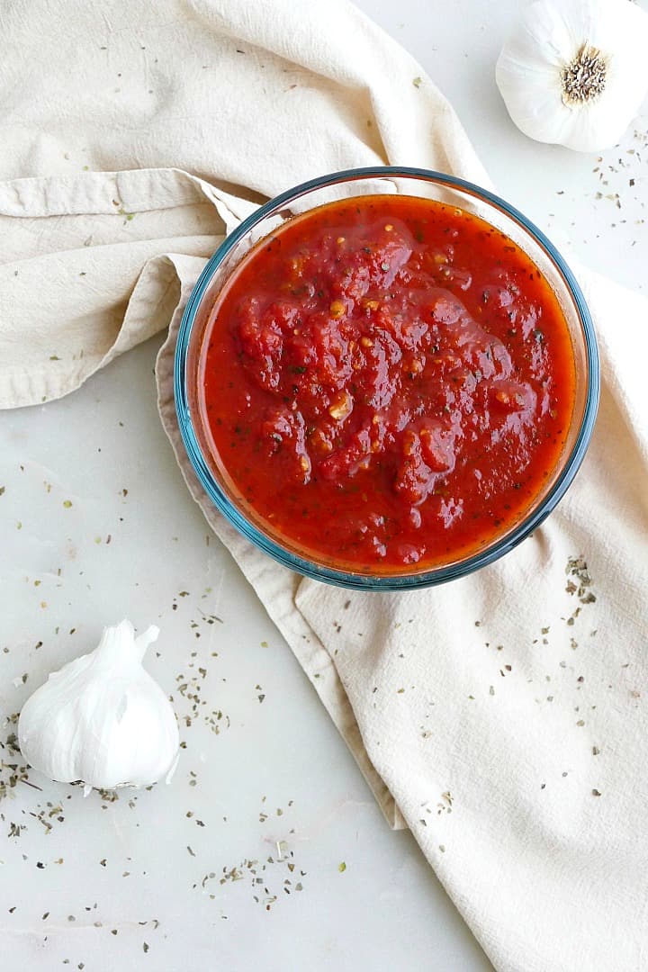 marinara sauce in a glass container on a napkin next to heads of garlic