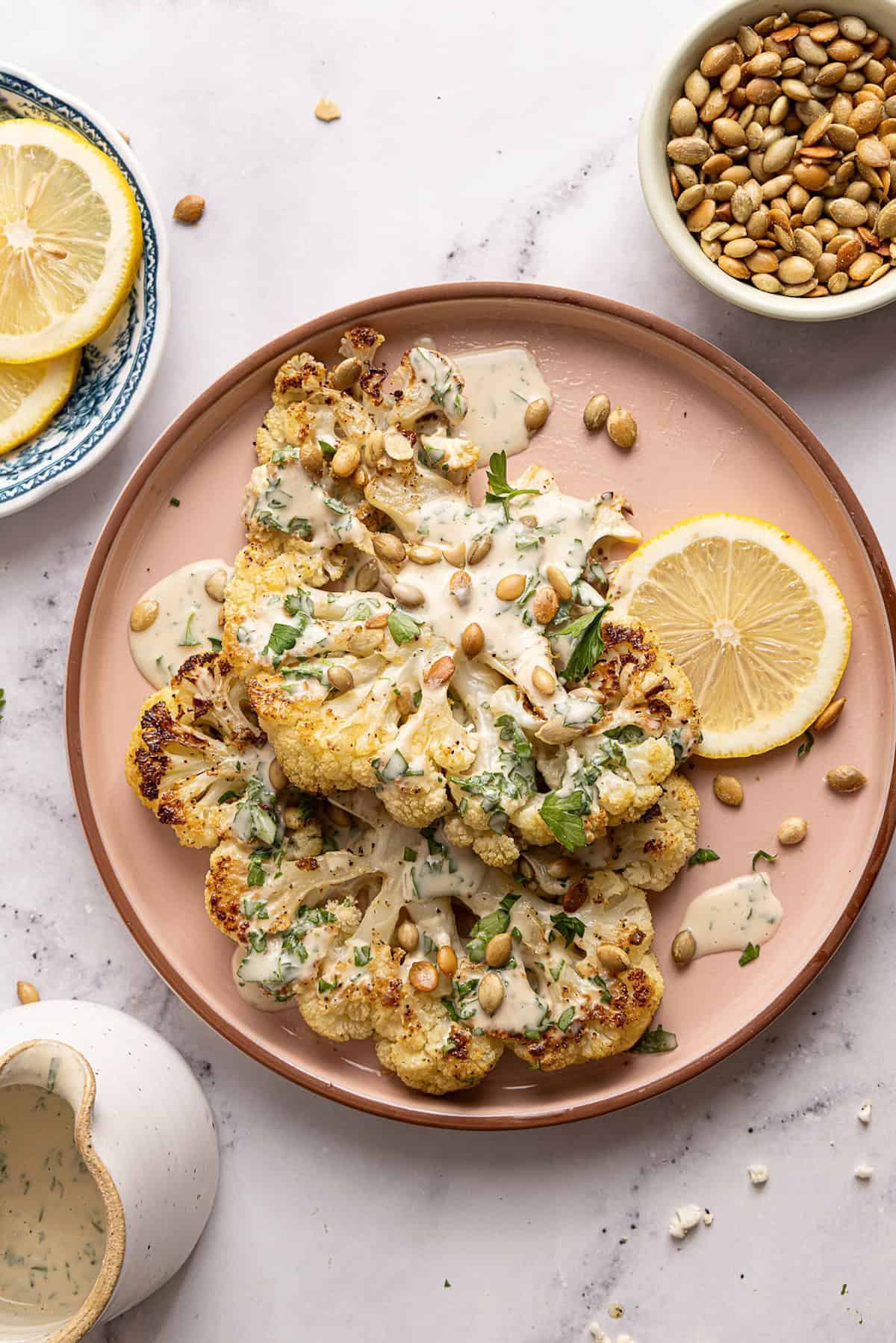 Cauliflower steaks with tahini sauce, pumpkin seeds, and parsley on a serving plate.