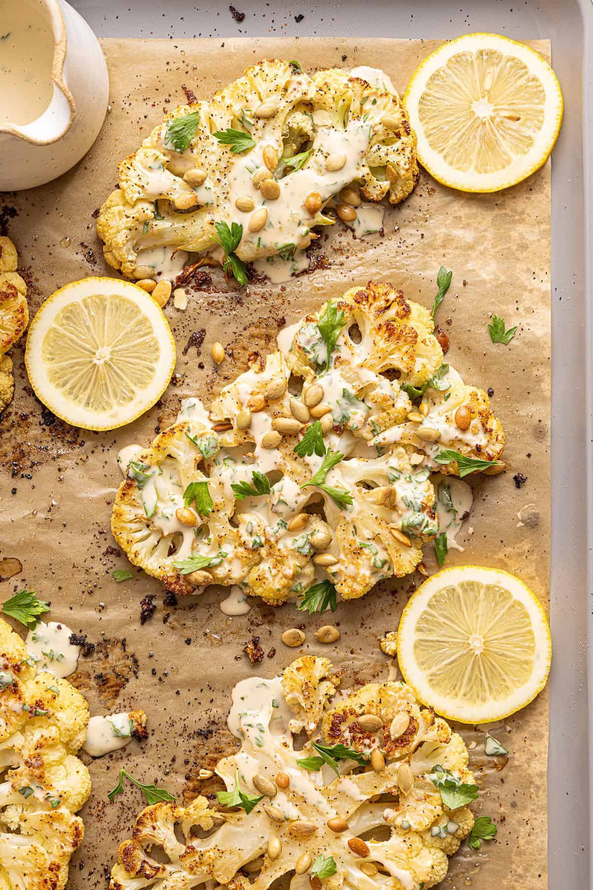 Cauliflower steaks with tahini and parsley on a baking sheet next to lemon slices.