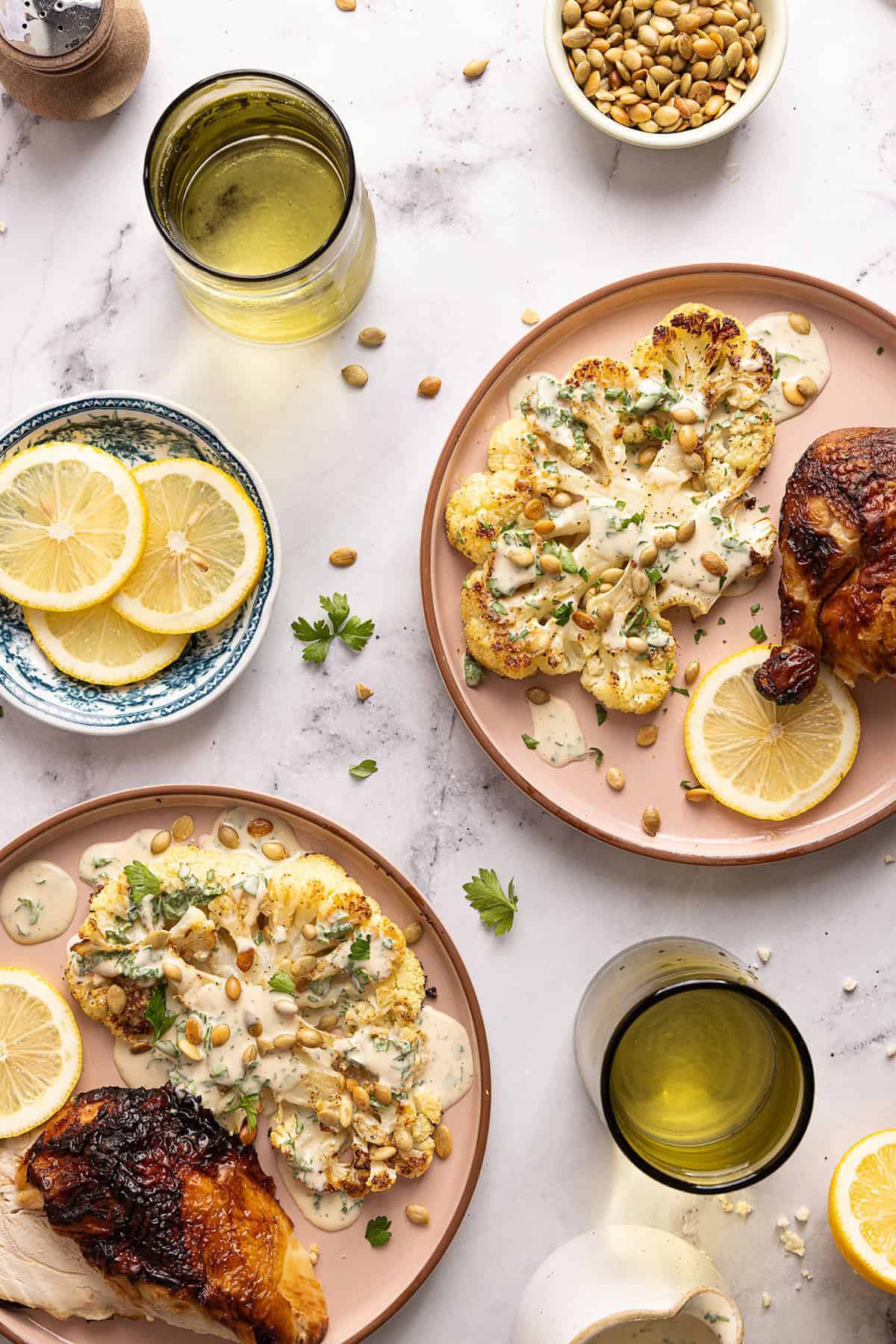 Two plates of a cauliflower steak recipe next to roasted chicken, lemons, and drinks.