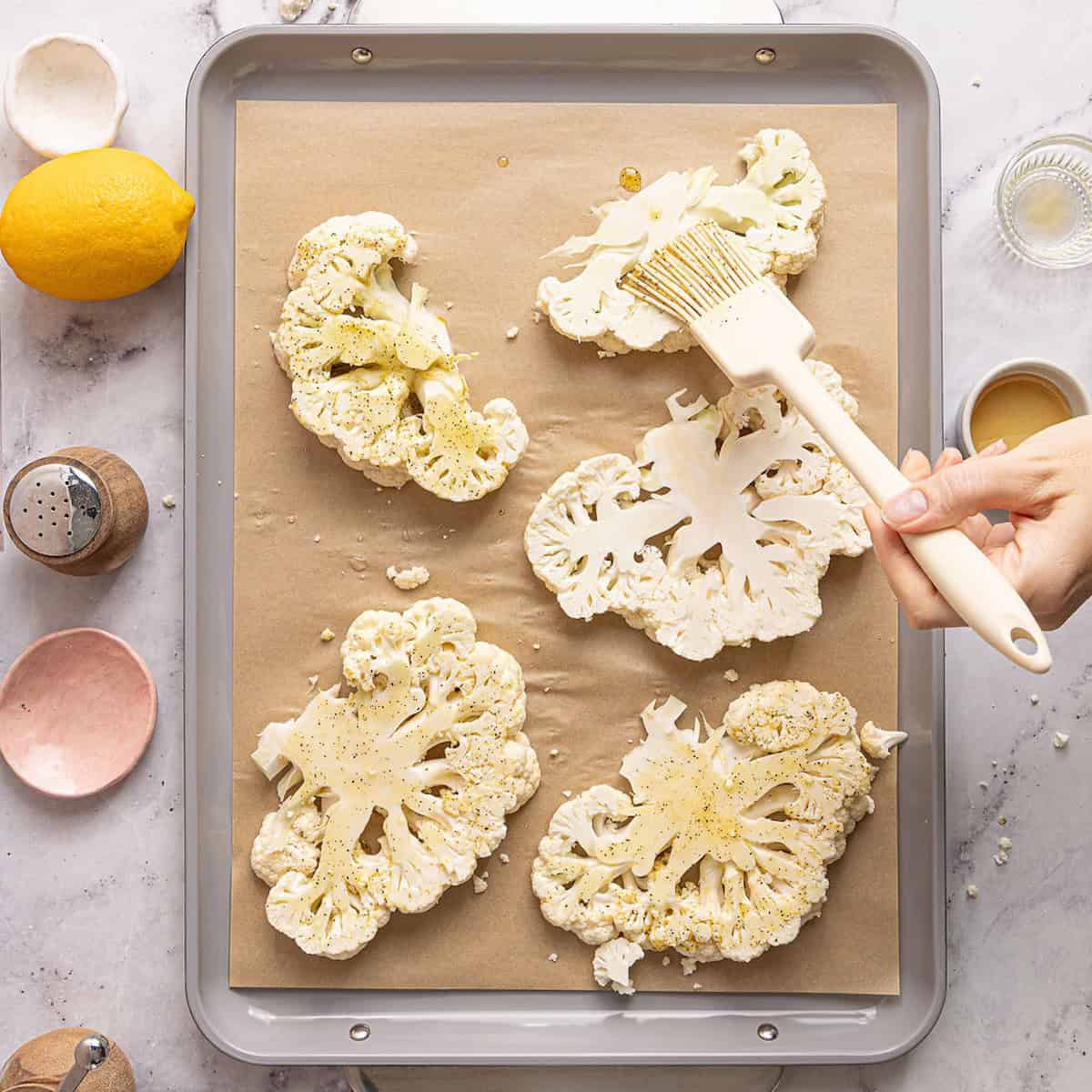 Woman brushing cauliflower "steaks" on a baking sheet with an olive oil spice mixture.