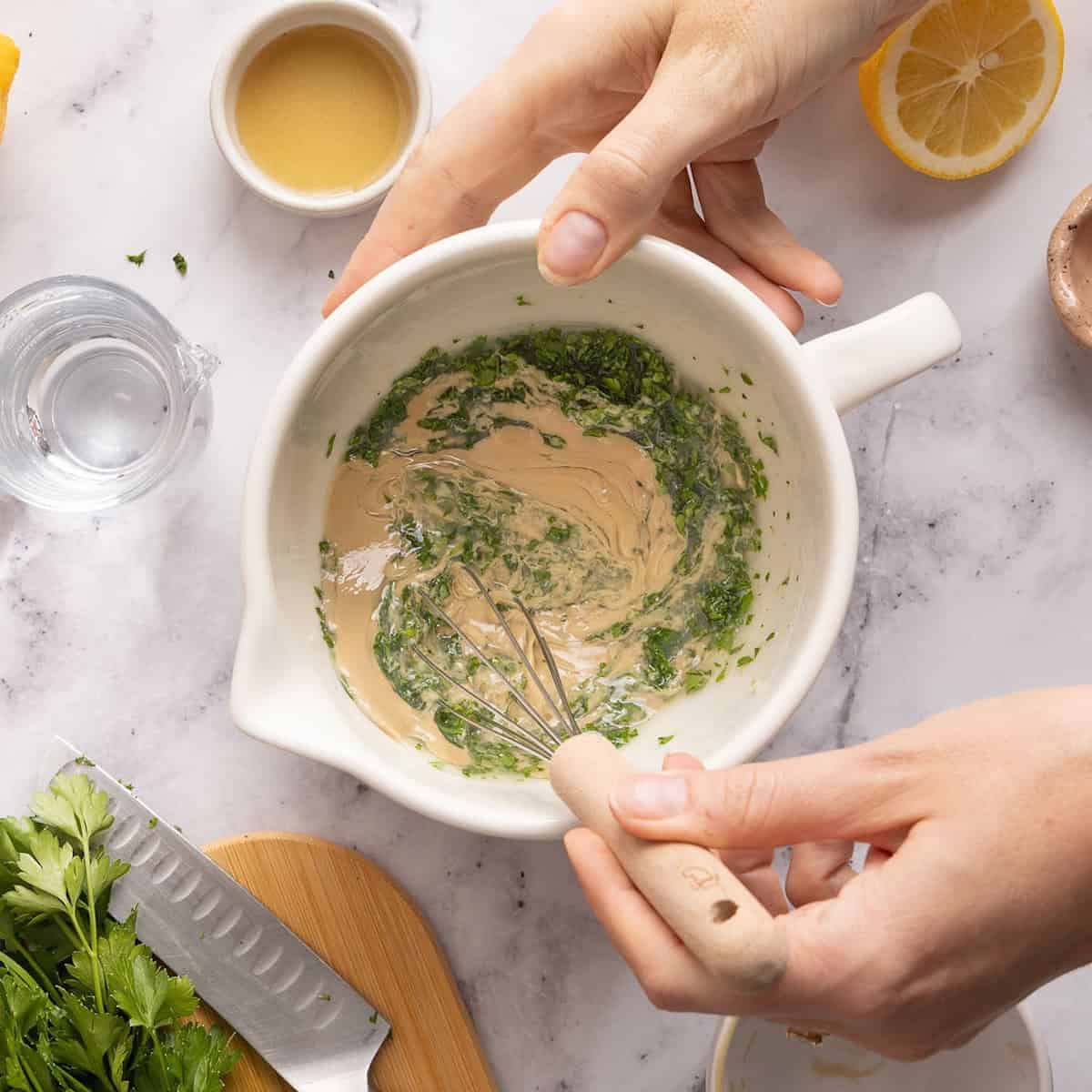 Woman whisking tahini with parsley, lemon juice, and water in a bowl.