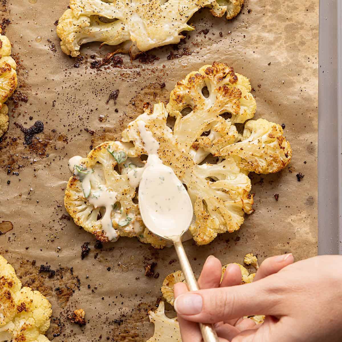 Woman spooning lemon parsley tahini sauce on roasted cauliflower on a baking sheet.