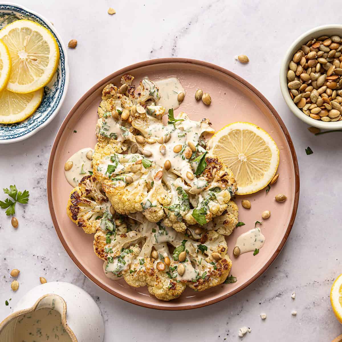 Cauliflower steaks with tahini, parsley, and pumpkin seeds on a serving plate with a lemon slice.