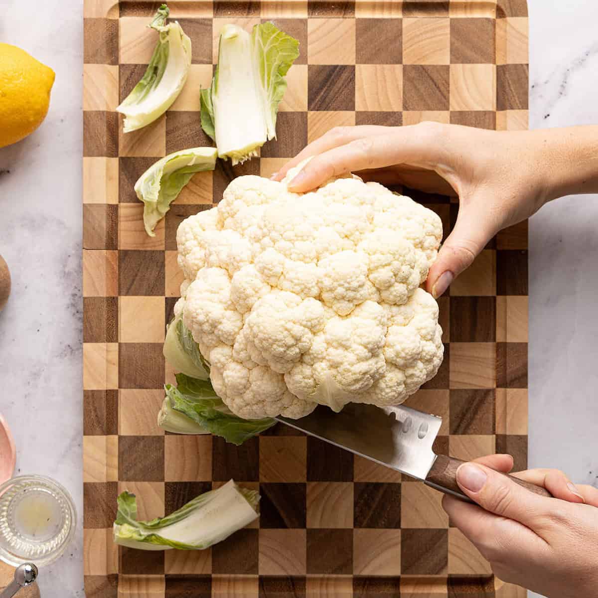 Woman trimming the leaves off of a head of cauliflower with a knife over a cutting board.