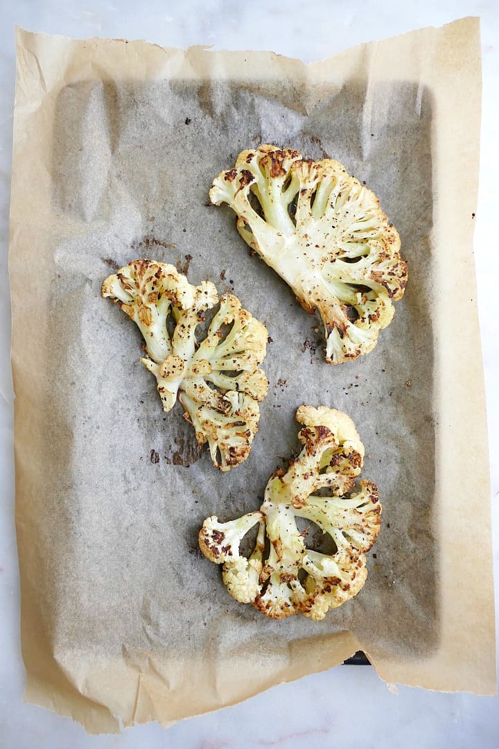 three roasted cauliflower steaks on a baking sheet lined with brown parchment paper