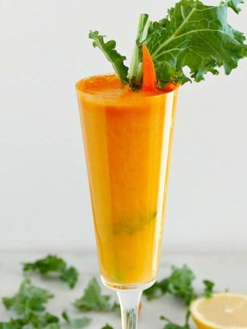square image of a champagne flute with a carrot juice cocktail garnished with kale