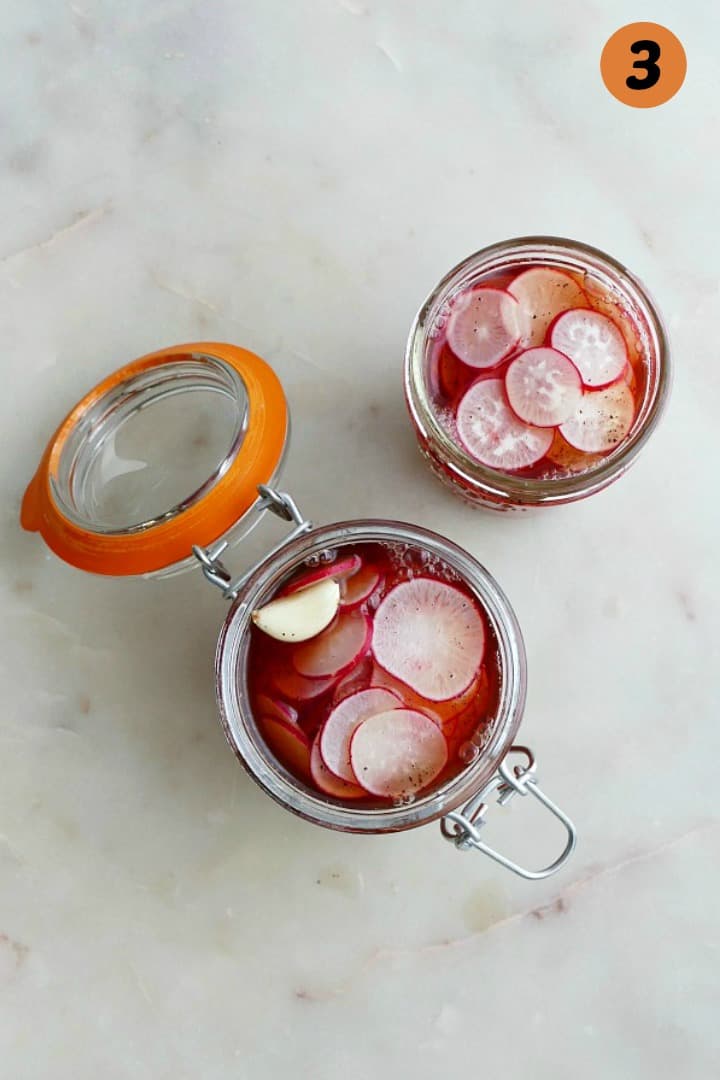 two glass jars filled with radishes and pickling brine on a white counter