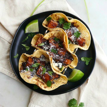 three tacos in corn tortillas on a black plate on top of a yellow napkin and counter