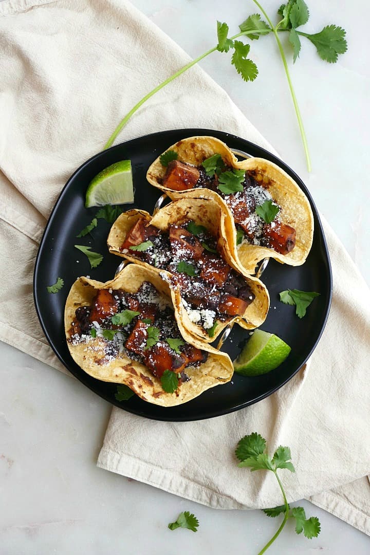 three tacos in corn tortillas on a black plate on top of a yellow napkin and counter