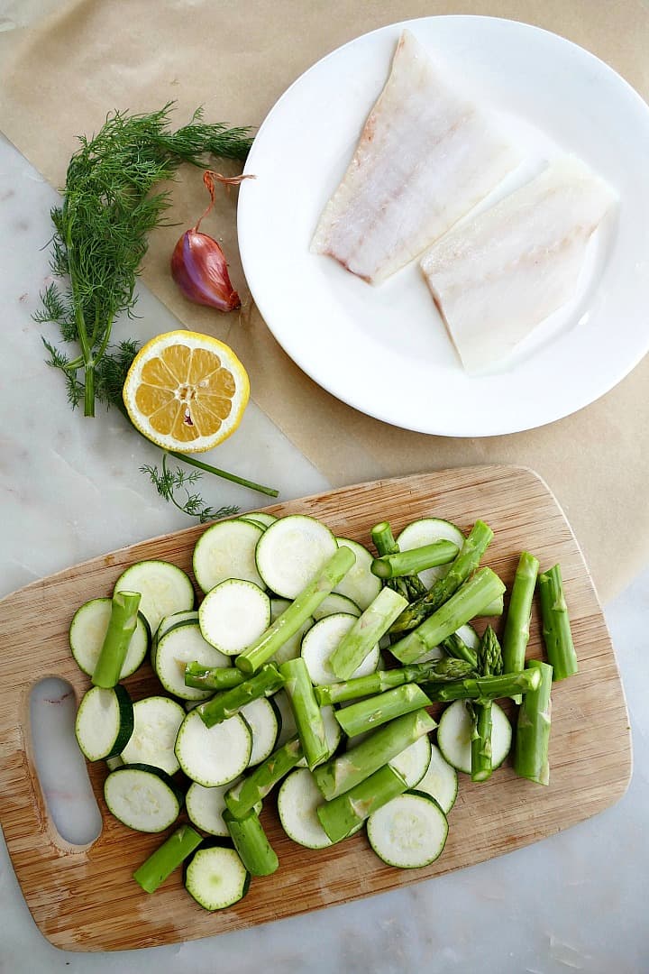 zucchini and asparagus on a cutting board next to cod fillets, dill, lemon, and shallot