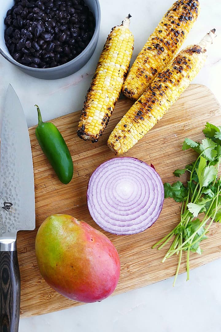 a cutting board with a mango, red onion, cilantro, jalapeno, and grilled corn
