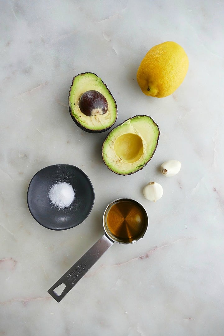 avocado, lemon, garlic cloves, salt, and olive oil on a white counter