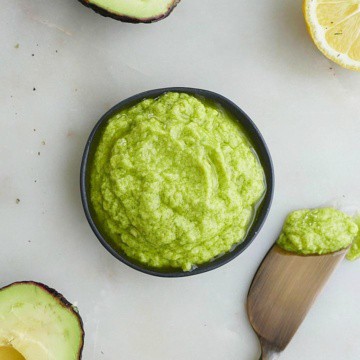 small black bowl of green avocado spread on a white counter surrounded by ingredients