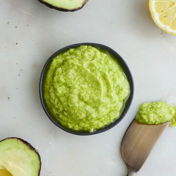 small black bowl of green avocado spread on a white counter surrounded by ingredients