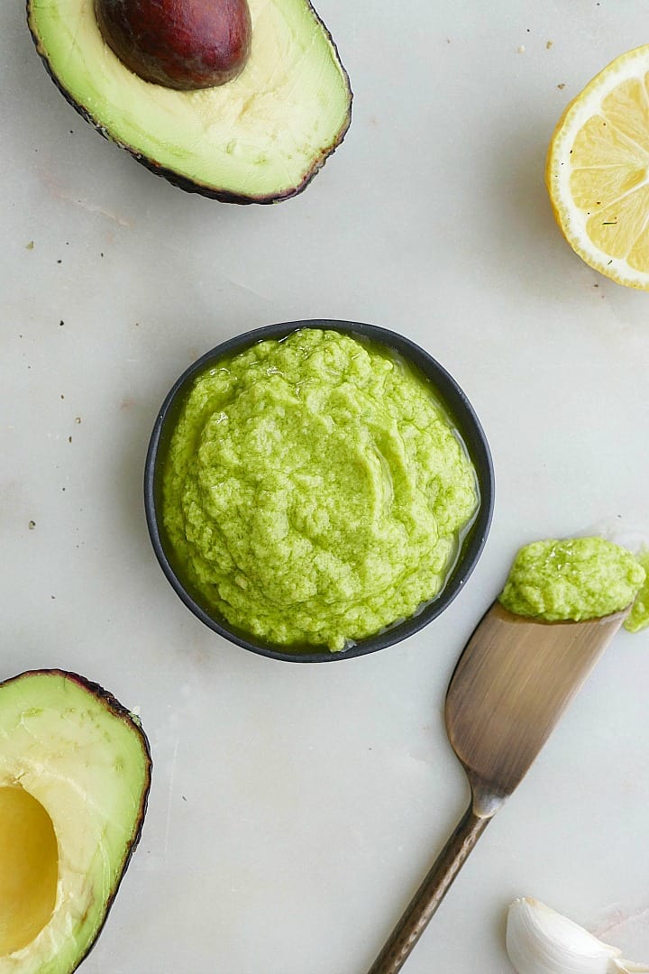 small black bowl of green avocado spread on a white counter surrounded by ingredients