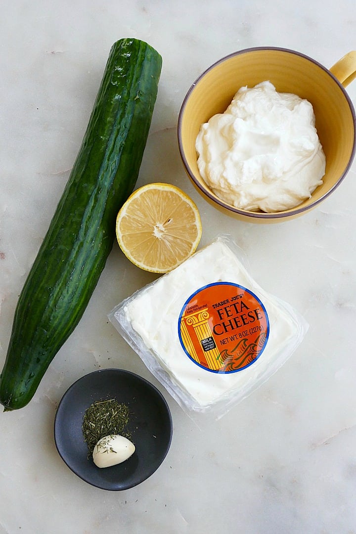 cucumber, feta block, lemon, garlic and herbs, and yogurt on a white counter
