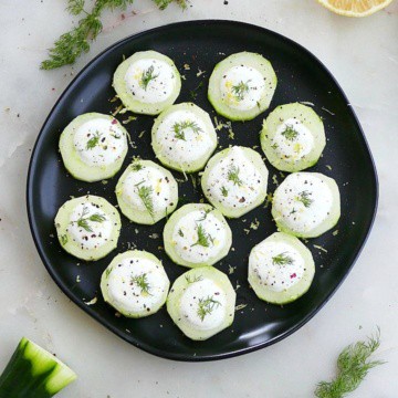 cucumber canapes on a black plate on a counter surrounded by dill