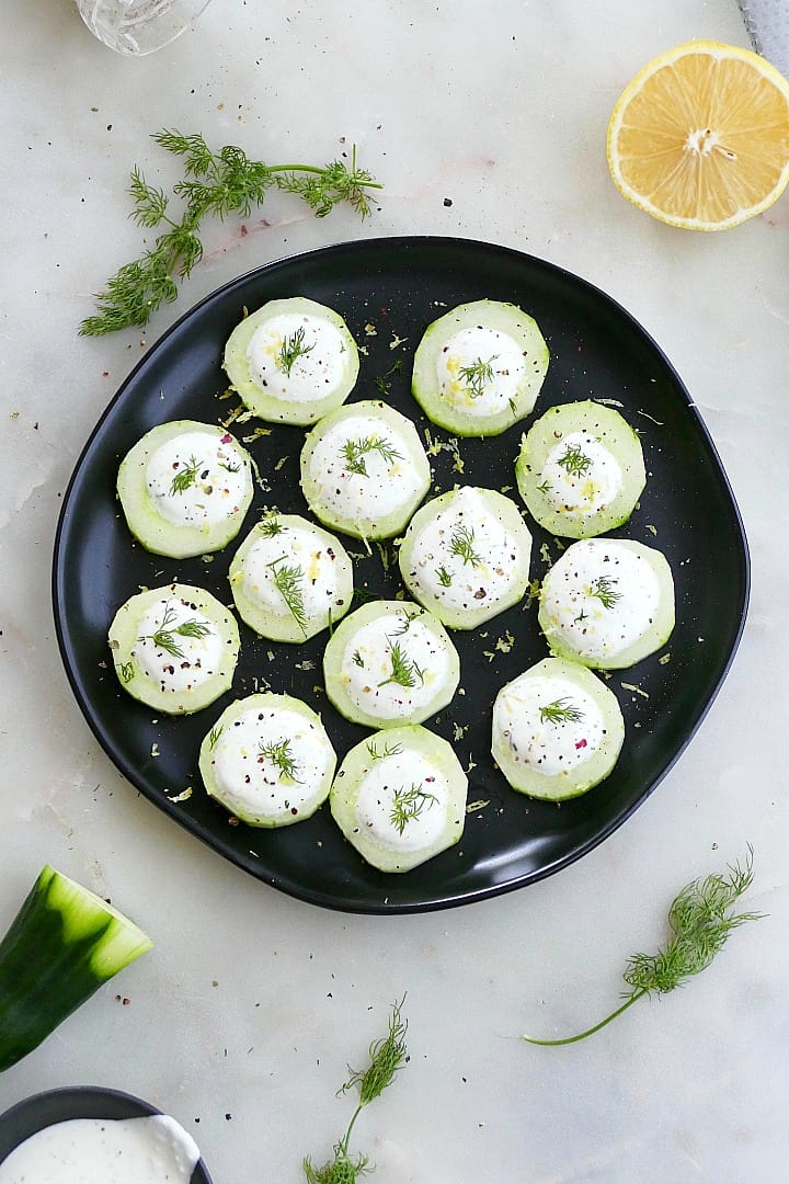 cucumber canapes on a black plate on a counter surrounded by dill