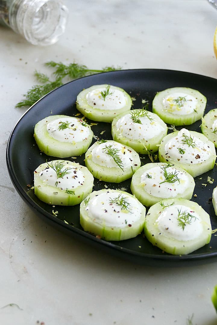 side view of cucumber canapes on a black plate topped with fresh dill