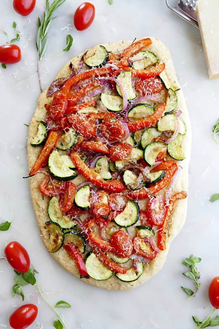 mediterranean roasted veggie flatbread on a white counter surrounded by ingredients and herbs