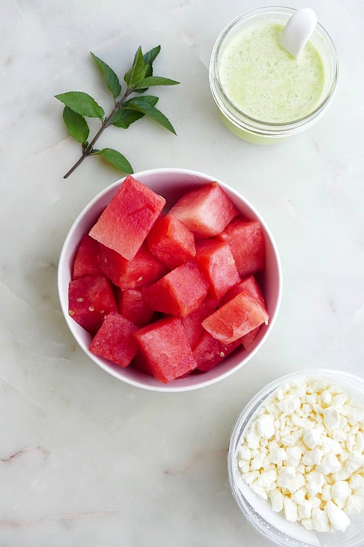 bowl of watermelon next to feta cheese, mint, and dressing on a counter