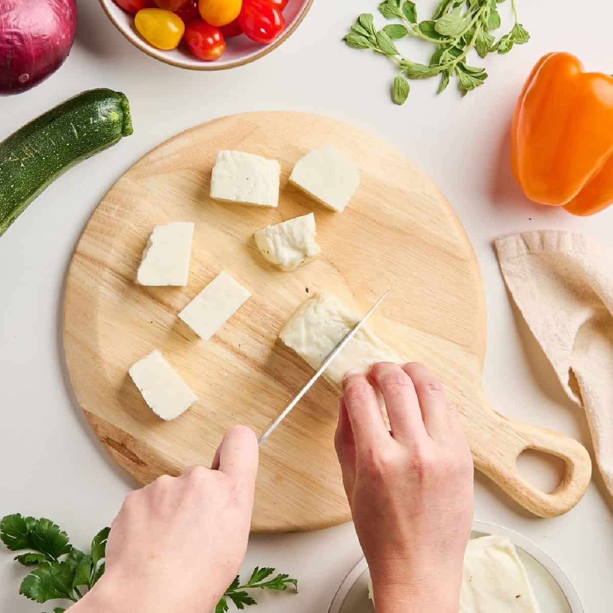 Woman cutting halloumi cheese into squares for skewers on a cutting board.