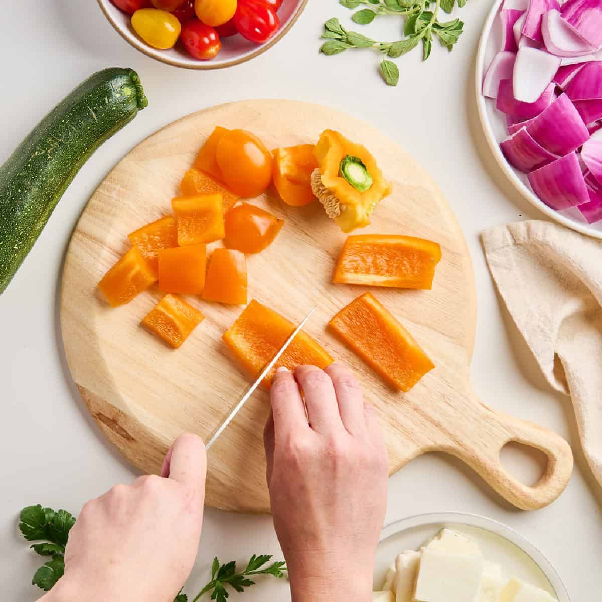 Woman cutting orange bell pepper into squares for skewers on a cutting board.