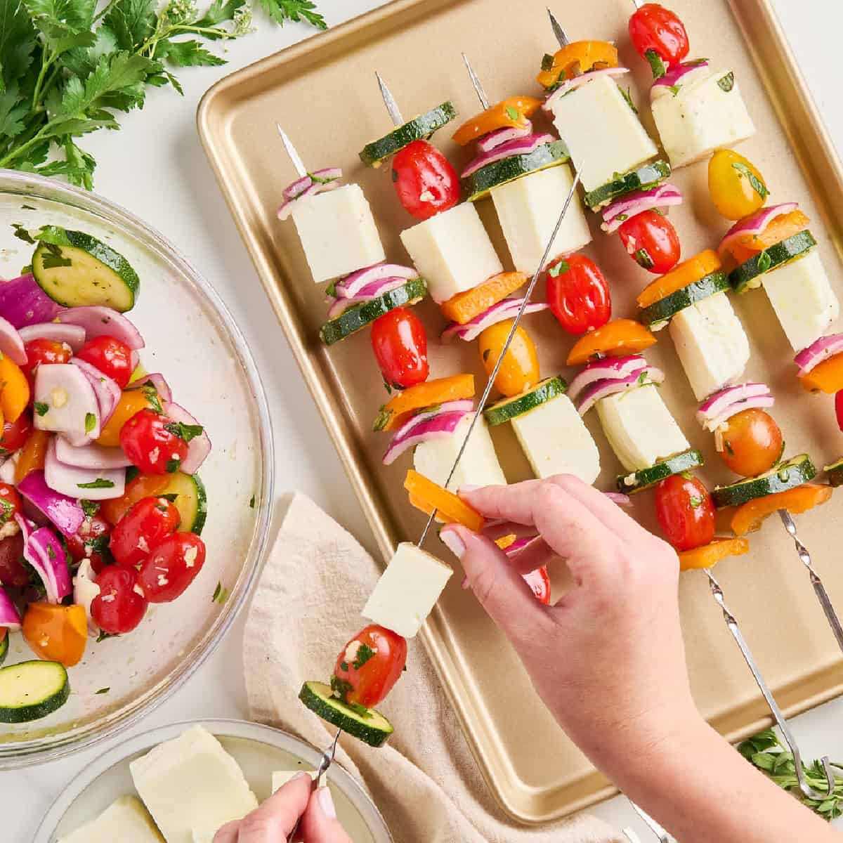 Woman putting bell pepper on a metal skewer with vegetables and halloumi.