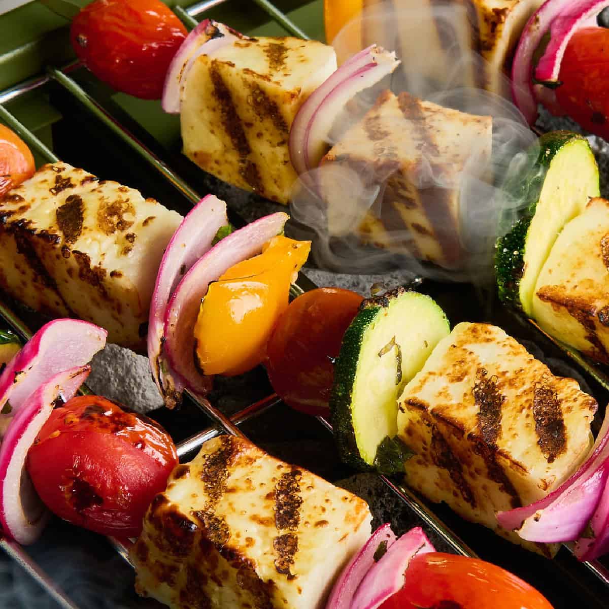 Vegetable skewers with halloumi on metal skewers cooking on grill grates until lightly charred.