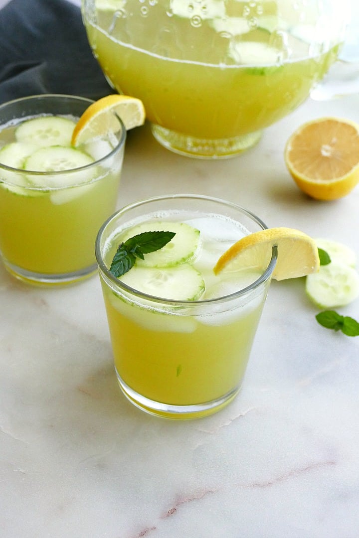 two glasses of cucumber mint lemonade in front of a pitcher on a counter