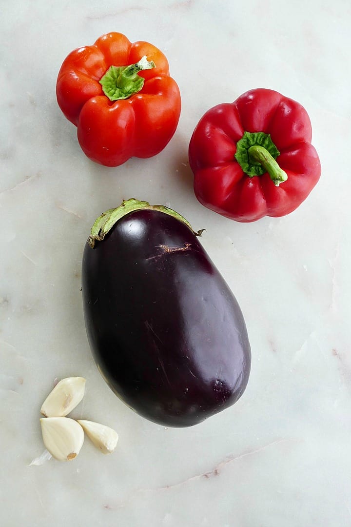 two red peppers, one large eggplant, and three garlic cloves spread out on a counter