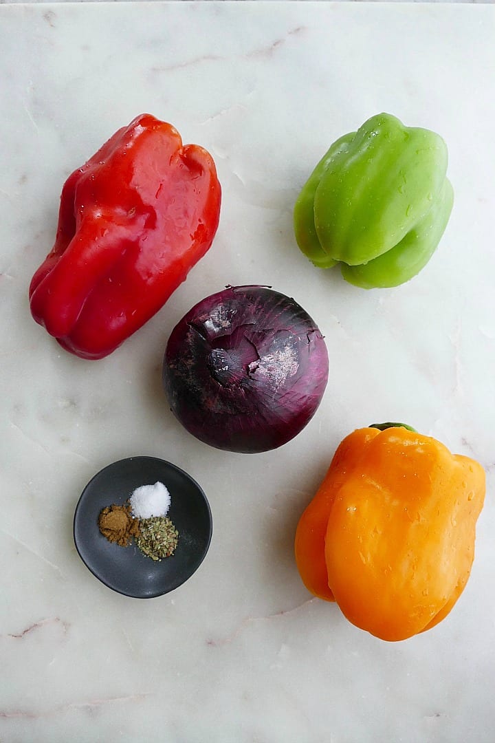 three bell peppers, a red onion, and a dish with seasonings on a counter