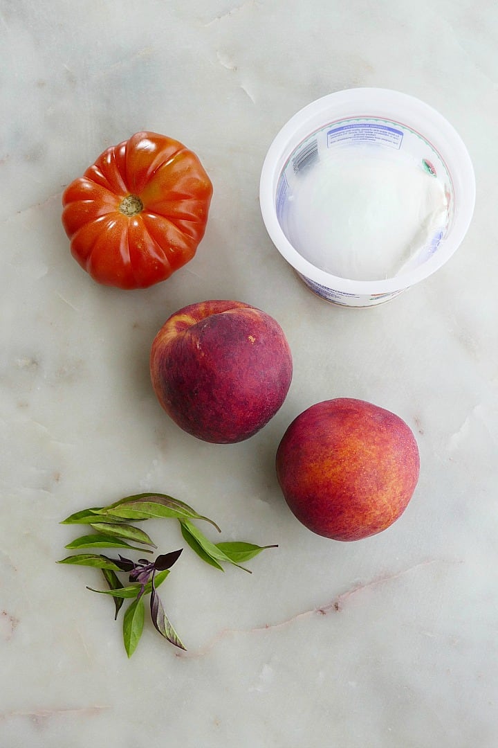 heirloom tomato, two peaches, ball of mozzarella, and basil leaves next to each other on a counter