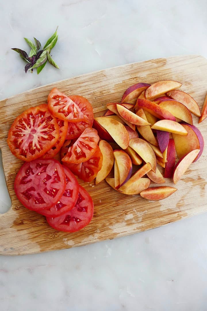 sliced tomatoes and peaches on a bamboo cutting board next to basil leaves