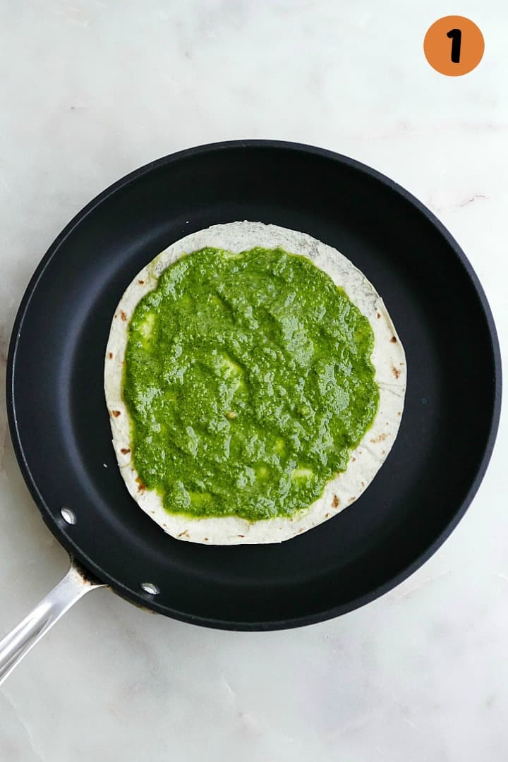 flour tortilla spread with green basil pesto on a black skillet on a counter