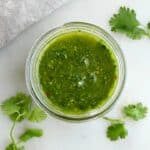 cilantro chimichurri sauce in a glass jar on a counter surrounded by herbs