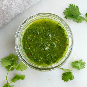 cilantro chimichurri sauce in a glass jar on a counter surrounded by herbs