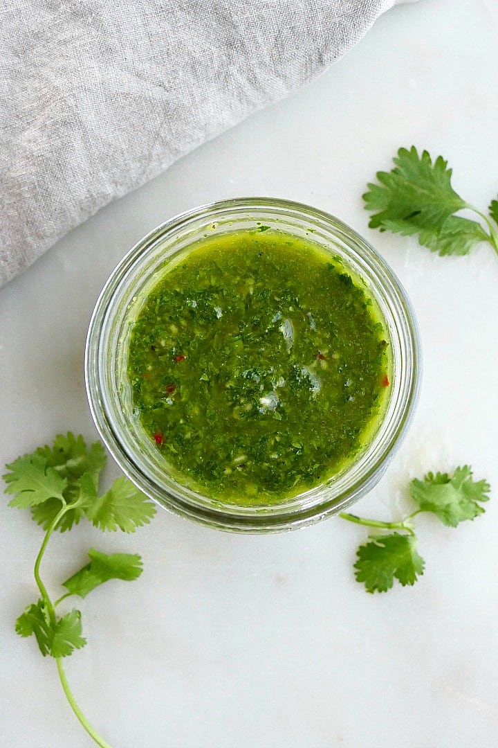 cilantro chimichurri sauce in a glass jar on a counter surrounded by herbs