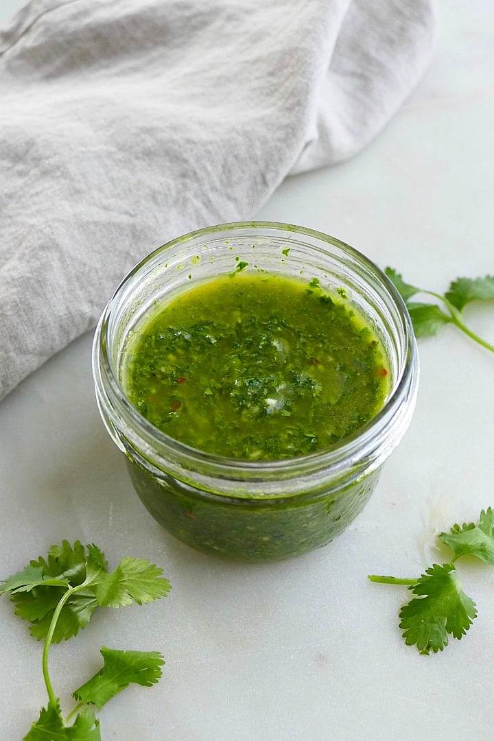 side view of cilantro chimichurri in a glass jar surrounded by herbs