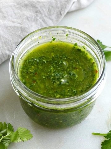 square image of cilantro chimichurri sauce in a jar surrounded by herbs
