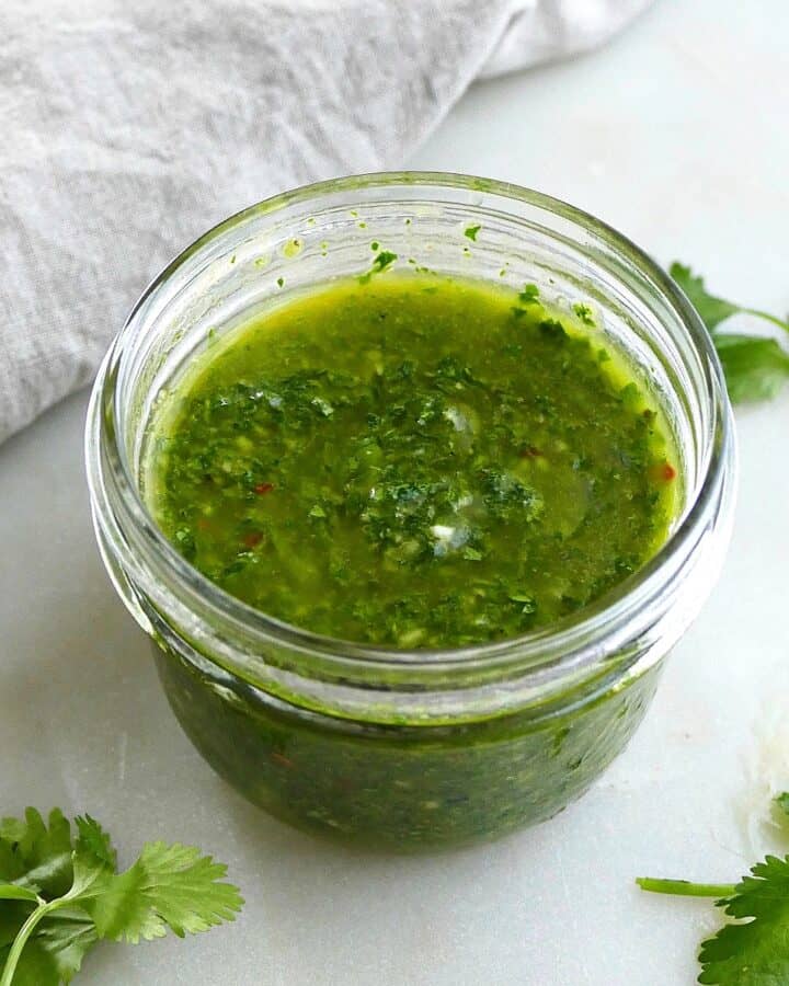 square image of cilantro chimichurri sauce in a jar surrounded by herbs