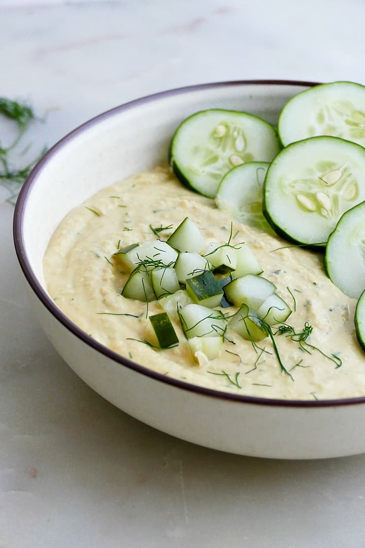 side view of dill pickle hummus garnished with fresh dill and cucumbers