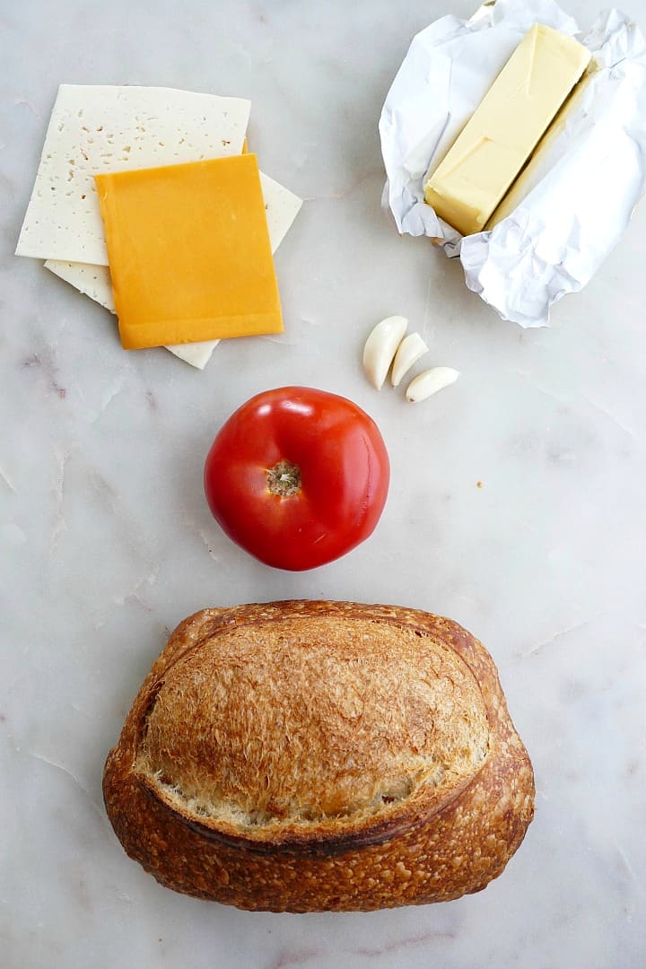 cheese slices, bread, garlic cloves, tomato, and butter spread out on a counter