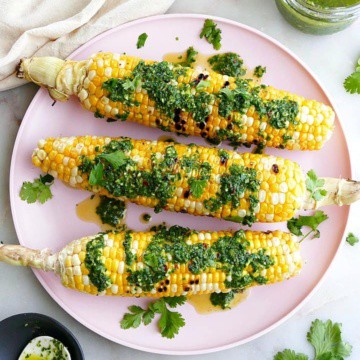 three grilled sweet corn cobs drizzled with chimichurri sauce on a serving platter