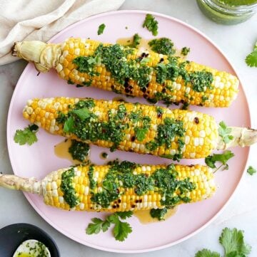 three grilled sweet corn cobs drizzled with chimichurri sauce on a serving platter