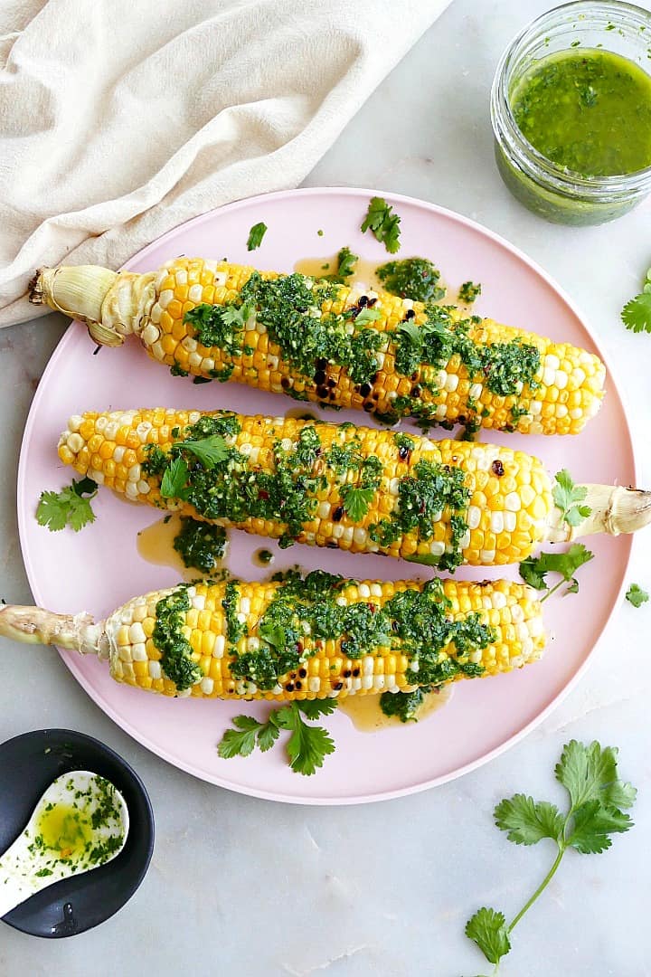 three grilled sweet corn cobs drizzled with chimichurri sauce on a serving platter