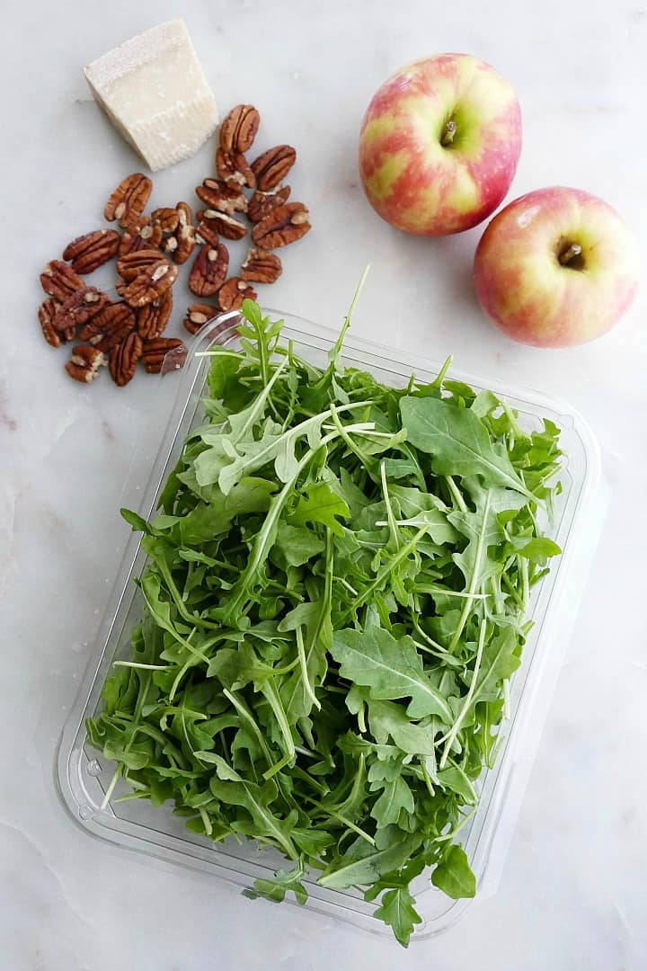 pecans, a block of parmesan, two apples, and arugula spread out on a counter