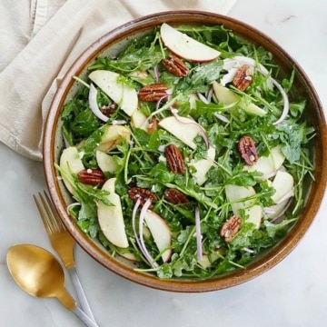 arugula apple salad in a large serving bowl next to gold and silver utensils